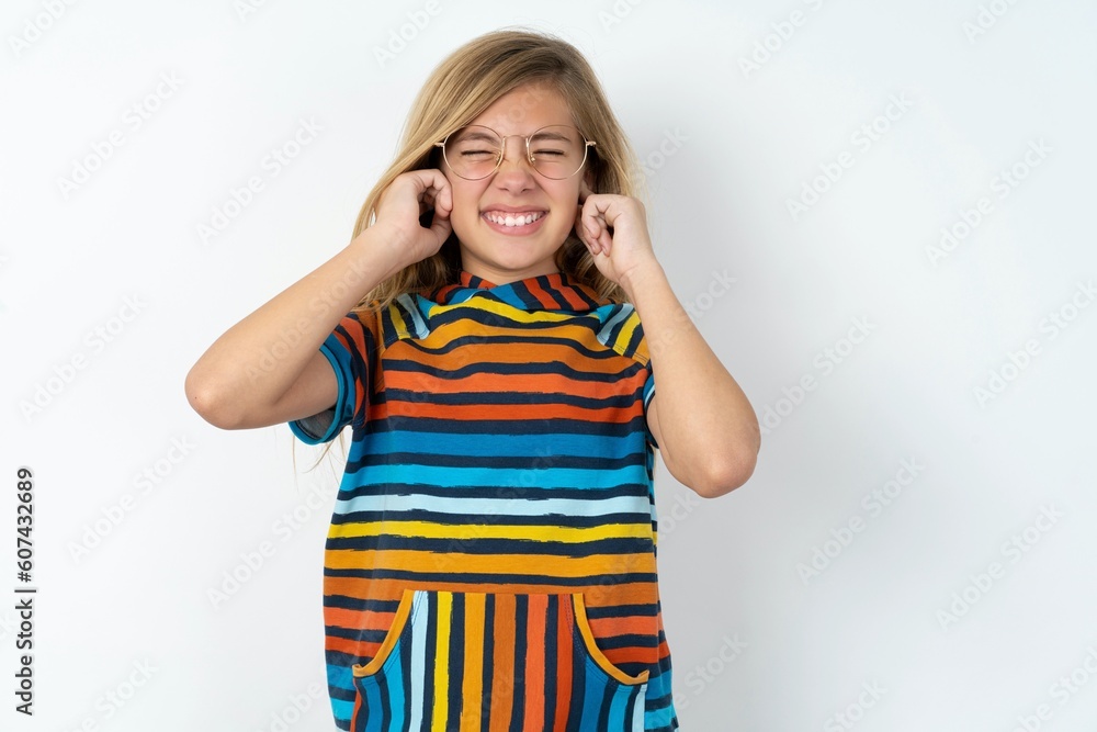 Stop making this annoying sound! Unhappy stressed out beautiful caucasian teen girl wearing striped T-shirt over white wall making worry face, plugging ears with fingers, irritated with loud noise. Stop making this annoying sound! Unhappy stressed out beautiful caucasian teen girl wearing striped T-shirt over white wall making worry face, plugging ears with fingers, irritated with loud noise.