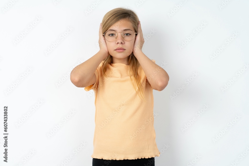 Frustrated beautiful caucasian teen girl wearing orange T-shirt over white wall plugging ears with hands does not wanting to listen hard rock, noise or loud music. Frustrated beautiful caucasian teen girl wearing orange T-shirt over white wall plugging ears with hands does not wanting to listen hard rock, noise or loud music.