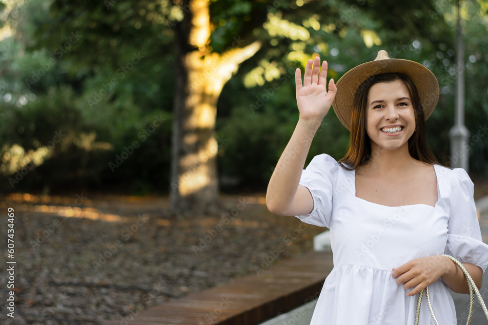 Naklejka premium Charming woman, smiling with white teeth, waving her hand, looking straight into the camera. A woman in a white dress, a straw hat on the background of the street. Stylish summer look, mixed