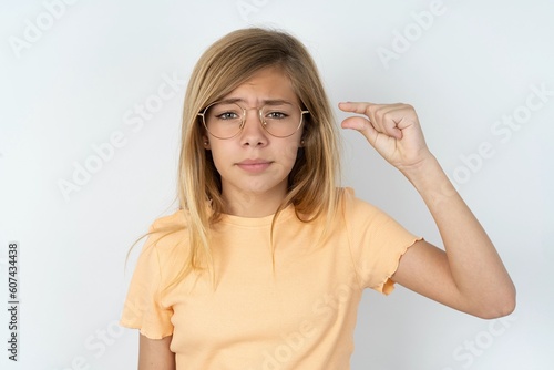 Upset beautiful caucasian teen girl wearing orange T-shirt over white wall shapes little gesture with hand demonstrates something very tiny small size. Not very much