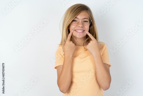 Strong healthy straight white teeth. Close up portrait of happy beautiful caucasian teen girl wearing orange T-shirt over white wall with beaming smile pointing on perfect clear white teeth.