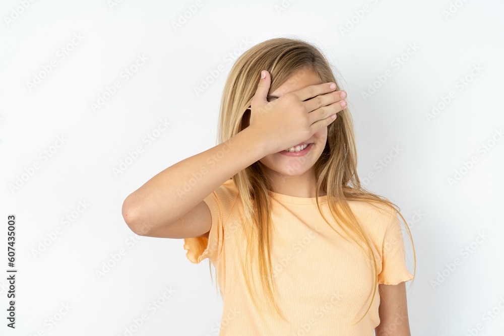 Happy beautiful caucasian teen girl wearing orange T-shirt over white wall closing eyes with hand going to see surprise prepared by friend standing and smiling in anticipation for something wonderful. Happy beautiful caucasian teen girl wearing orange T-shirt over white wall closing eyes with hand going to see surprise prepared by friend standing and smiling in anticipation for something wonderful.