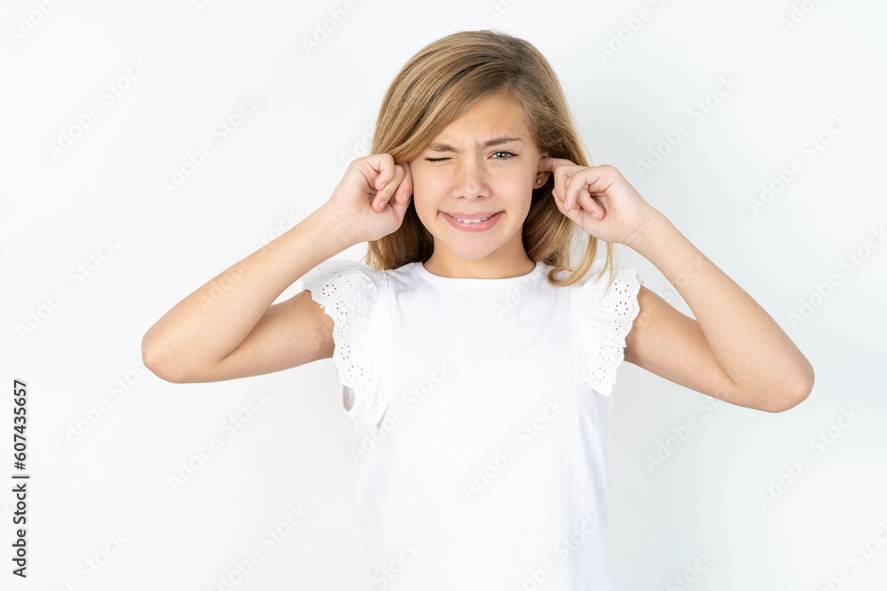 Happy beautiful caucasian teen girl wearing white T-shirt over white wall ignores loud music and plugs ears with fingers asks to turn off sound Happy beautiful caucasian teen girl wearing white T-shirt over white wall ignores loud music and plugs ears with fingers asks to turn off sound