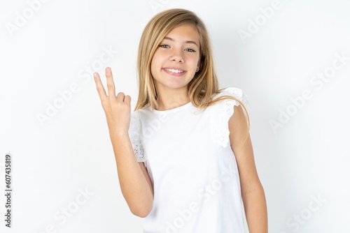beautiful caucasian teen girl wearing white T-shirt over white wall smiling and looking friendly, showing number two or second with hand forward, counting down