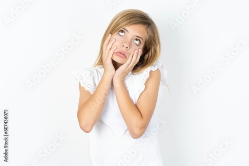 beautiful caucasian teen girl wearing white T-shirt over white wall keeps hands on cheeks has bored displeased expression. Stressed hopeless model