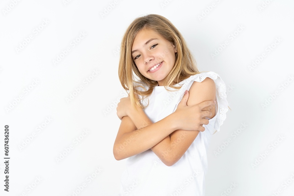 Charming pleased beautiful caucasian teen girl wearing white T-shirt over white wall embraces own body, pleasantly feels comfortable poses. Tenderness and self esteem concept Charming pleased beautiful caucasian teen girl wearing white T-shirt over white wall embraces own body, pleasantly feels comfortable poses. Tenderness and self esteem concept
