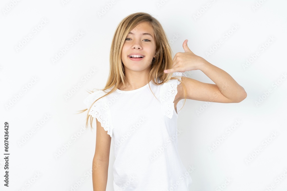 beautiful caucasian teen girl wearing white T-shirt over white wall makes phone gesture, says call me back again, has glad expression. beautiful caucasian teen girl wearing white T-shirt over white wall makes phone gesture, says call me back again, has glad expression.
