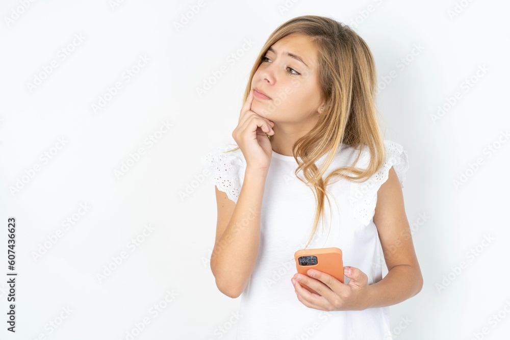 beautiful caucasian teen girl wearing white T-shirt over white wall thinks deeply about something, uses modern mobile phone, tries to made up good message, keeps index finger near lips. beautiful caucasian teen girl wearing white T-shirt over white wall thinks deeply about something, uses modern mobile phone, tries to made up good message, keeps index finger near lips.