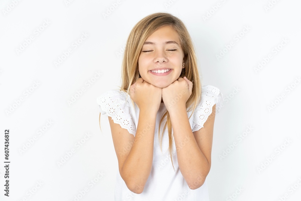 Cheerful beautiful caucasian teen girl wearing white T-shirt over white wall has shy satisfied expression, smiles broadly, shows white teeth, People emotions Cheerful beautiful caucasian teen girl wearing white T-shirt over white wall has shy satisfied expression, smiles broadly, shows white teeth, People emotions