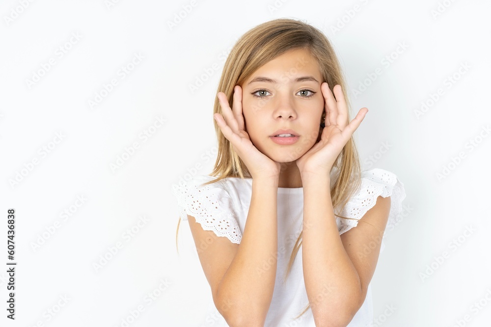 Happy beautiful caucasian teen girl wearing white T-shirt over white wall touches both cheeks gently, has tender smile, shows white teeth, gazes positively straightly at camera, Happy beautiful caucasian teen girl wearing white T-shirt over white wall touches both cheeks gently, has tender smile, shows white teeth, gazes positively straightly at camera,