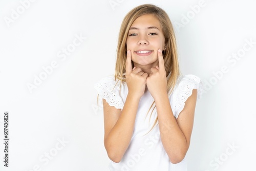 Happy beautiful caucasian teen girl wearing white T-shirt over white wall with toothy smile  keeps index fingers near mouth  fingers pointing and forcing cheerful smile