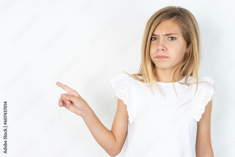 Serious beautiful caucasian teen girl wearing white T-shirt over white wall smirks face points away on copy space shows something unpleasant. Look at this advertisement. Big price concept Serious beautiful caucasian teen girl wearing white T-shirt over white wall smirks face points away on copy space shows something unpleasant. Look at this advertisement. Big price concept