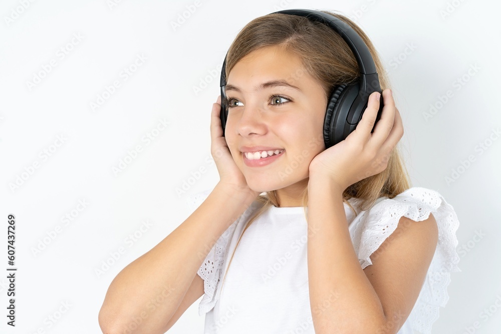 beautiful caucasian teen girl wearing white T-shirt over white wall wears stereo headphones listens music concentrated aside. People hobby lifestyle concept beautiful caucasian teen girl wearing white T-shirt over white wall wears stereo headphones listens music concentrated aside. People hobby lifestyle concept