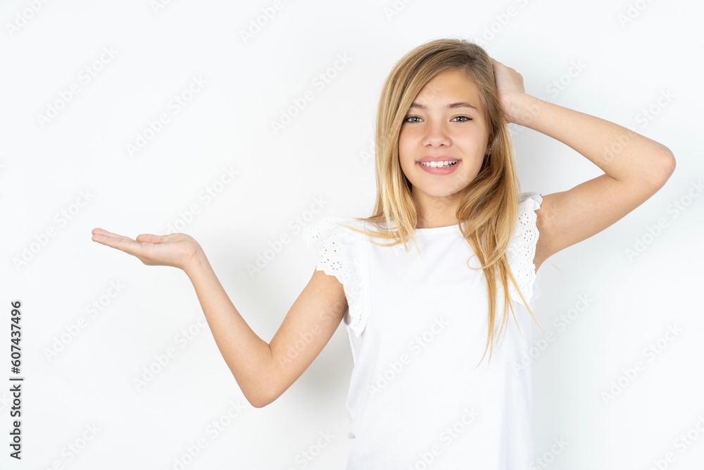 Fototapeta premium Shocked amazed surprised beautiful caucasian teen girl wearing white T-shirt over white wall hold hand offering proposition