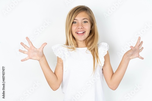 Delighted positive beautiful caucasian teen girl wearing white T-shirt over white wall opens mouth and arms palms up after having great result