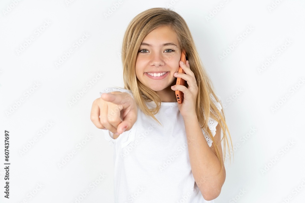 Positive beautiful caucasian teen girl wearing white T-shirt over white wall indicates directly at camera has telephone conversation smiles broadly enjoys talking long hours. You join me Positive beautiful caucasian teen girl wearing white T-shirt over white wall indicates directly at camera has telephone conversation smiles broadly enjoys talking long hours. You join me
