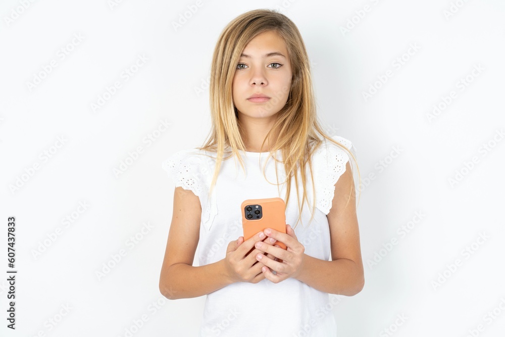Portrait of serious confident beautiful caucasian teen girl wearing white T-shirt over white wall holding phone in two hands Portrait of serious confident beautiful caucasian teen girl wearing white T-shirt over white wall holding phone in two hands