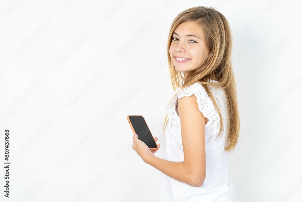 Rear view photo portrait of beautiful caucasian teen girl wearing white T-shirt over white wall using smartphone smiling Rear view photo portrait of beautiful caucasian teen girl wearing white T-shirt over white wall using smartphone smiling