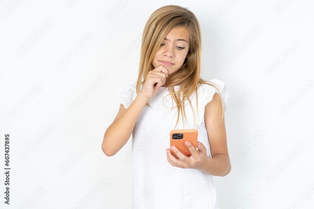 Thoughtful happy beautiful caucasian teen girl wearing white T-shirt over white wall hold look phone, copyspace Thoughtful happy beautiful caucasian teen girl wearing white T-shirt over white wall hold look phone, copyspace