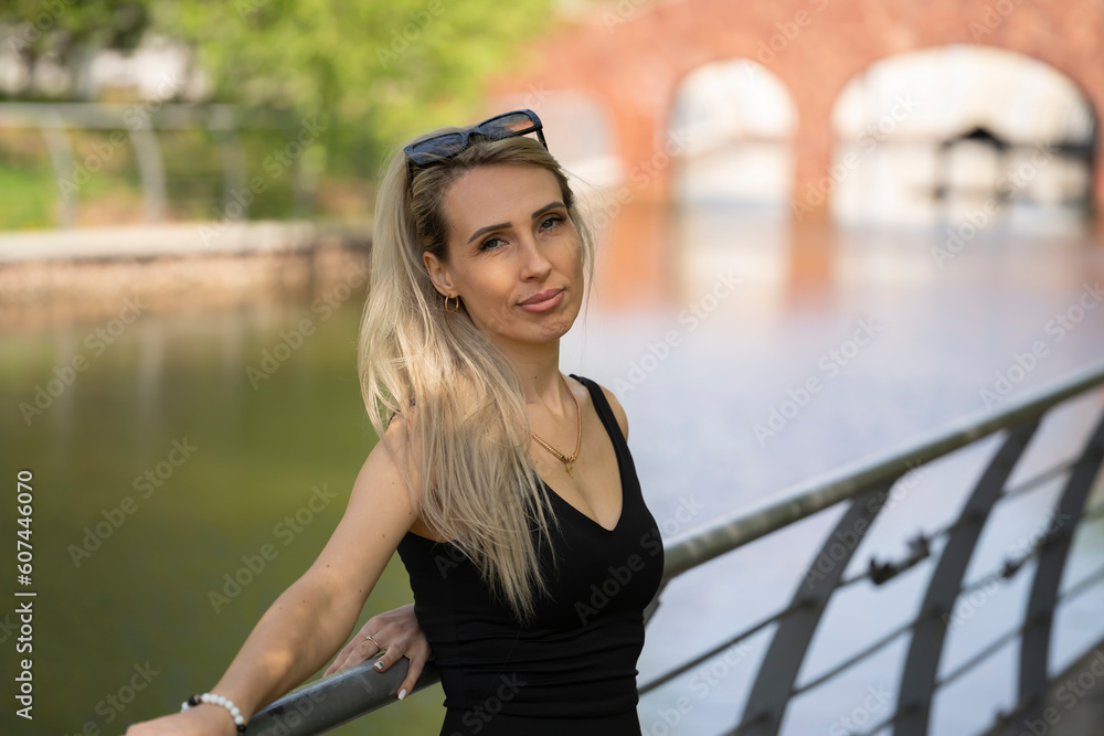 A young blonde woman in sunglasses stands on the bank of a pond in a city park. The girl looks into the camera. High quality photo