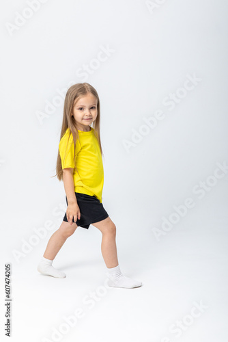 Calm and self-confident cute blonde kid girl in summer casual clothes, passes us looking at camera over white background. Stylish comfortable everyday fashion for children concept