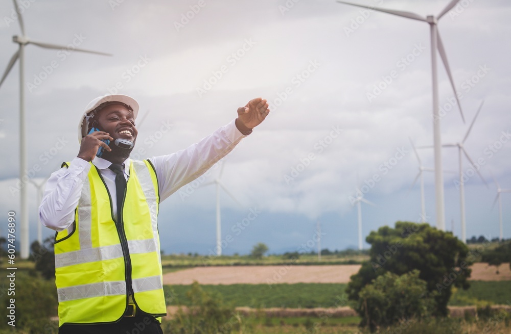 African engineer man stands using smartphone front the wind turbines generating electricity power station. .Concept of sustainability development by alternative energy
