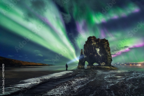 Fotografie Aurora borealis above Hvitserkur rock formation, A man with a lantern (headlamp)