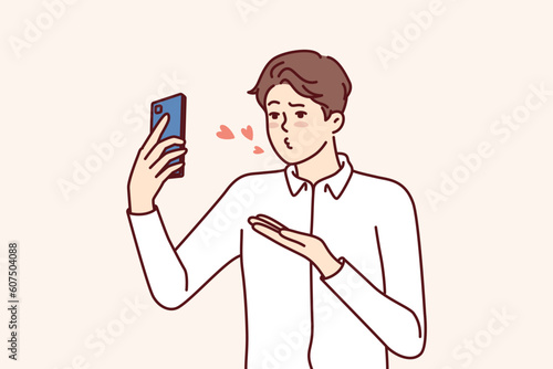 Man with mobile phone sends air kiss to interlocutor during video call to girlfriend who has left on business trip. Guy with smartphone records video of valentine wishing to kiss bride from distance