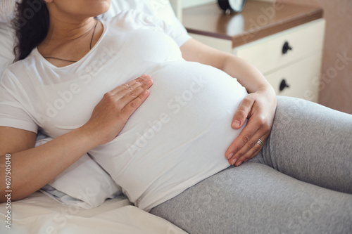 Close-up. Belly of a pregnant woman, expectant mother in white t-shirt gently caressing her abdomen, relaxing, lying on the bed at home interior. Happy pregnancy, procreation, maternity leave concept