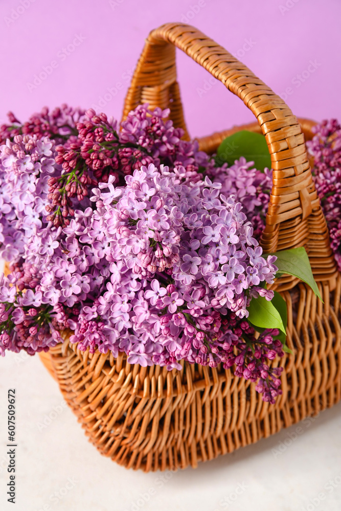 Fototapeta premium Basket with beautiful lilac flowers on light table against color background, closeup