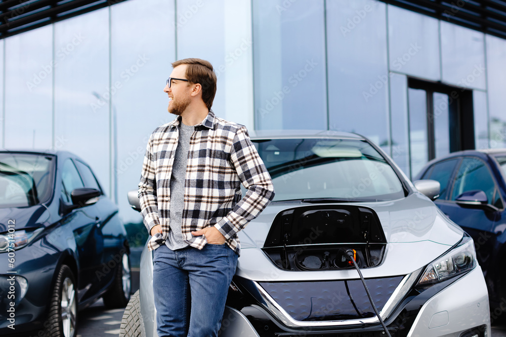 A young man stands next to his new electric car and rests while the car is charging A young man stands next to his new electric car and rests while the car is charging