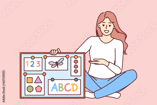 Woman english teacher in elementary school with magnetic board with alphabet and geometric shapes. Girl is sitting on floor explaining rules of english language to young children in accessible way.