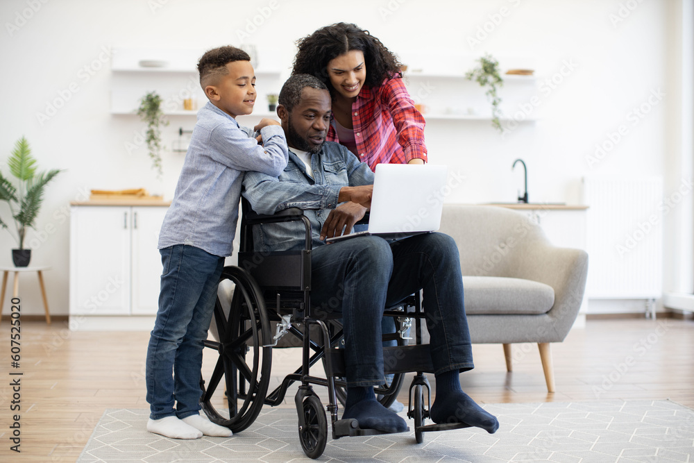 Cheerful multiracial man using wheelchair holding laptop on knees while pretty woman and funny boy standing behind. Loving family of three choosing movie for watching together on Sunday at home. Cheerful multiracial man using wheelchair holding laptop on knees while pretty woman and funny boy standing behind. Loving family of three choosing movie for watching together on Sunday at home.