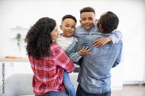 Back view of multicultural spouses holding preschool boys in arms while having break from playing board games. Friendly family planning after-dinner walk before leaving cozy home on sunny day.