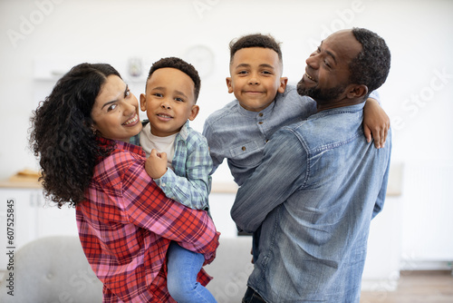 Portrait of smiling multiethnic man and woman with cute kids in arms looking over shoulder in apartment interior. Delighted parents and cute sons displaying affection for each member of loving family.