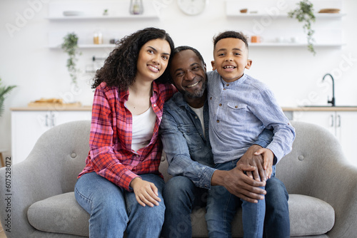 Portrait of cheerful multicultural family wearing jeans posing on sofa in modern lounge of studio apartment. Playful little boy hugging mom and dad while getting positive energy from parents' care.