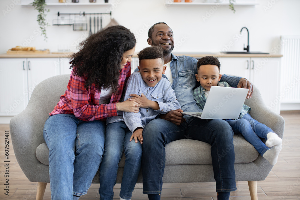 Affectionate african american mom tickling one of cute sons while happy father showing video to another boy. Cheerful parents and children using wireless laptop while having fun at home. Affectionate african american mom tickling one of cute sons while happy father showing video to another boy. Cheerful parents and children using wireless laptop while having fun at home.