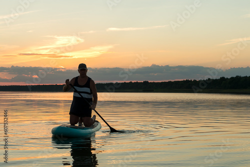A man on his knees on a SUP board with an oar at sunset swims in the calm water of the lake against the backdrop of a pink sky reflection in the water.