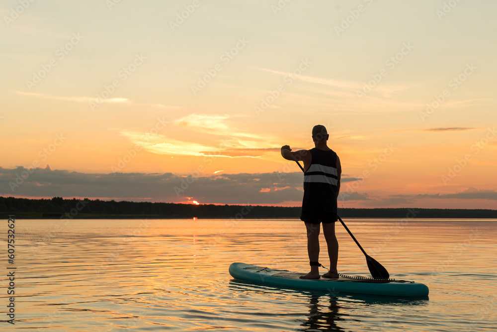 A man on a SUP board with a paddle at sunset against a pink-orange sky in the water of the lake.