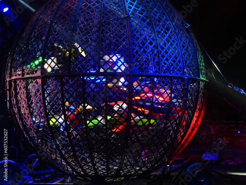 Group of five bikers performing maneuvers inside a spherical cage, known as the globe of death, at night, illuminated by artificial lights and colored LEDs