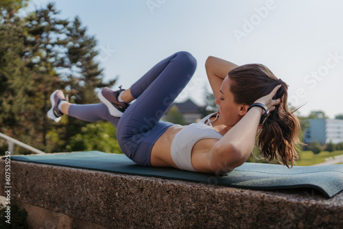 Fototapeta Naklejka Na Ścianę i Meble -  Side view of young sporty woman doing bicycle crunches on gym mat in the city.