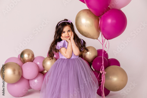 Cute smiling little girl in a lilac princess dress posing with air balloons isolated on white background. Kids Birthday party celebration concept. Happy Birthday banner with copy space. Studio shot.