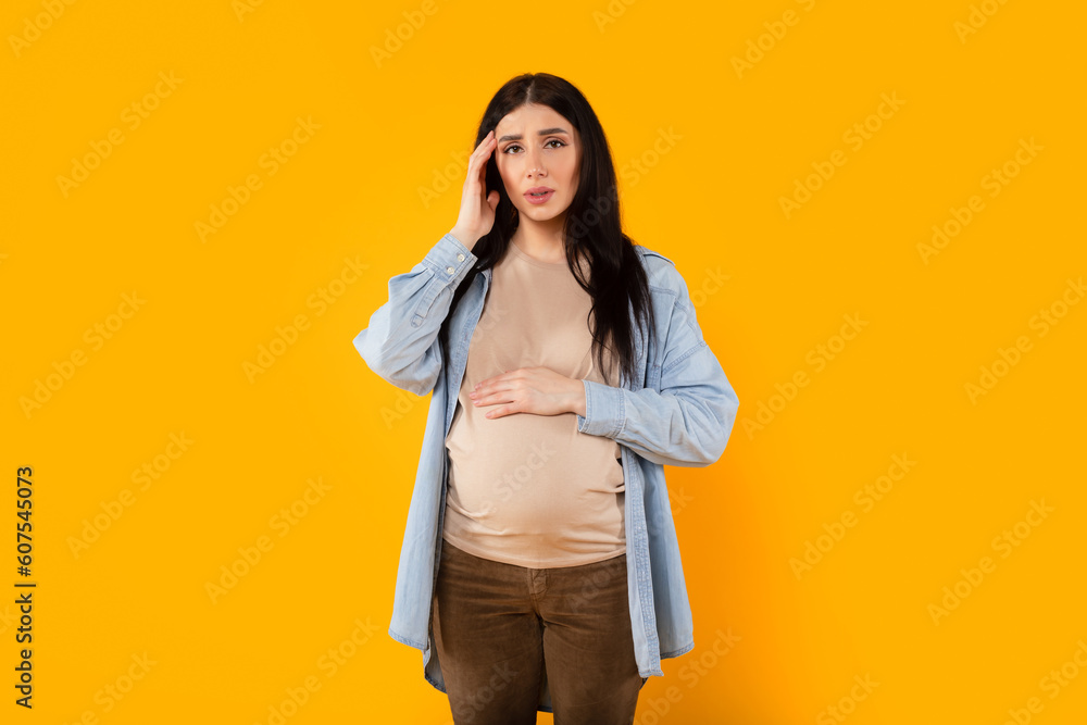 Sick pregnant woman suffering headache while standing over yellow studio background, expectant lady touching temples