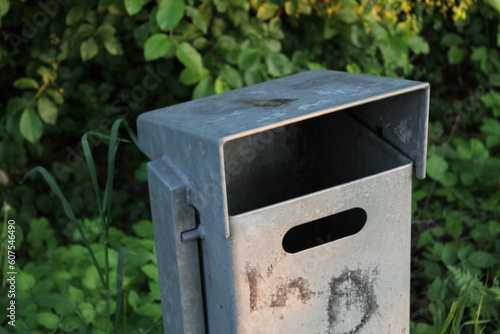 Metal trash can in the park - waste disposal with leaves in the background.