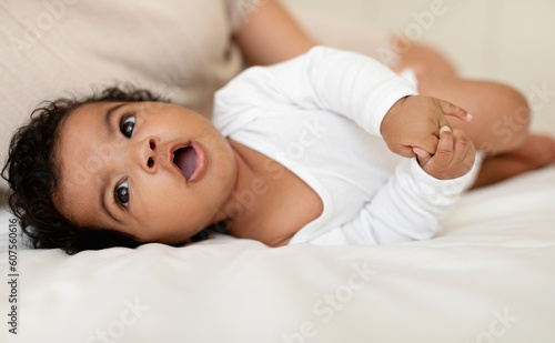 Happy sweet black little baby with open mouth lies on white bed with mom in bedroom