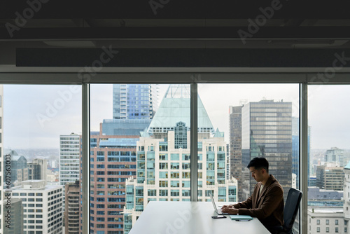 Young adult Asian business man executive working on laptop at corporate office. Professional Japanese businessman manager using computer technology sitting at table with big city view from window.