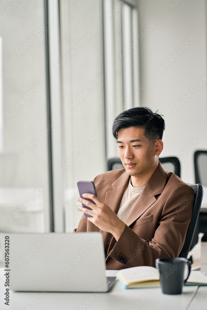 Young busy Asian Japanese business man worker executive holding cellphone device using mobile cell phone looking at smartphone checking applications working in corporate office with laptop, vertical. Young busy Asian Japanese business man worker executive holding cellphone device using mobile cell phone looking at smartphone checking applications working in corporate office with laptop, vertical.