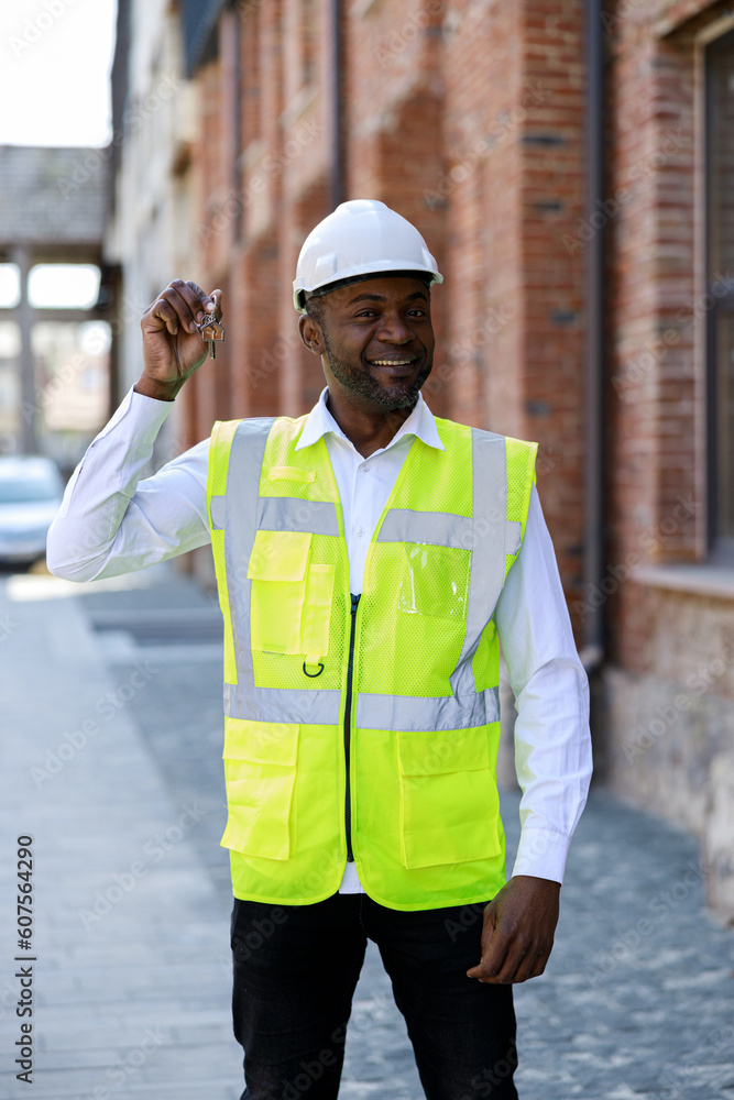Portrait of smiling construction engineer holding keys from new home. Happy african american worker wearing safety helmet looking at camera with happy face outdoors. Portrait of smiling construction engineer holding keys from new home. Happy african american worker wearing safety helmet looking at camera with happy face outdoors.