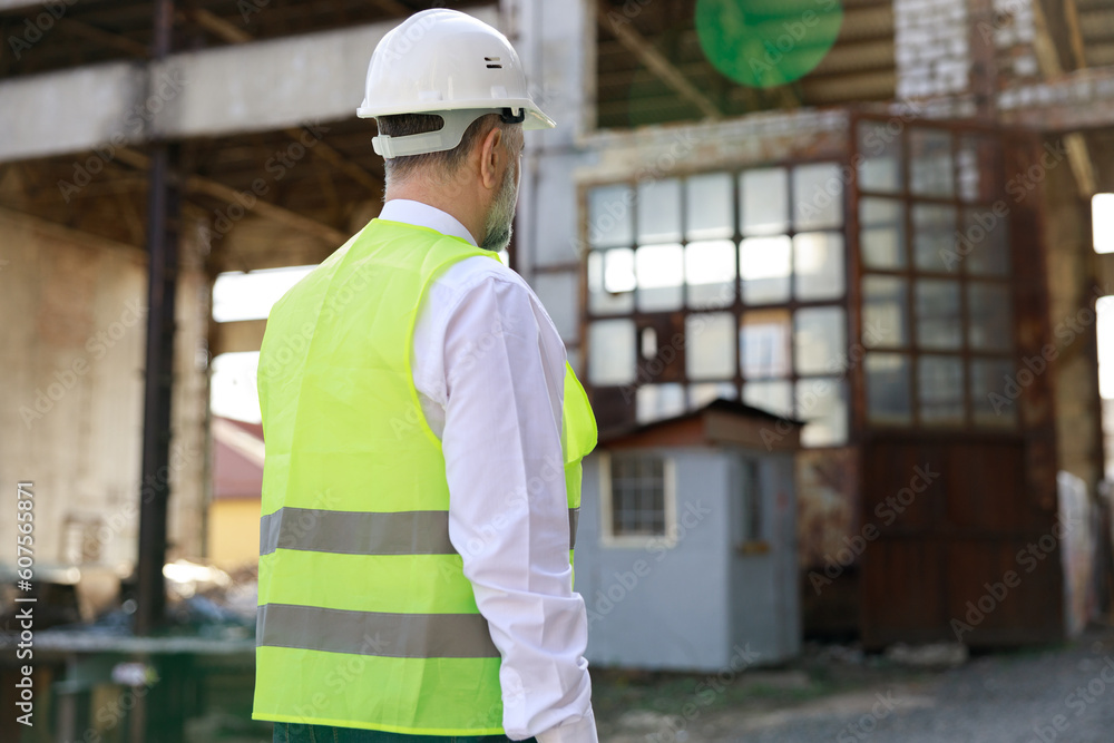 Back view of caucasian male builder in hardhat walking along wall of unfinished building. Competent architect examining and controlling working process at construction site. Back view of caucasian male builder in hardhat walking along wall of unfinished building. Competent architect examining and controlling working process at construction site.