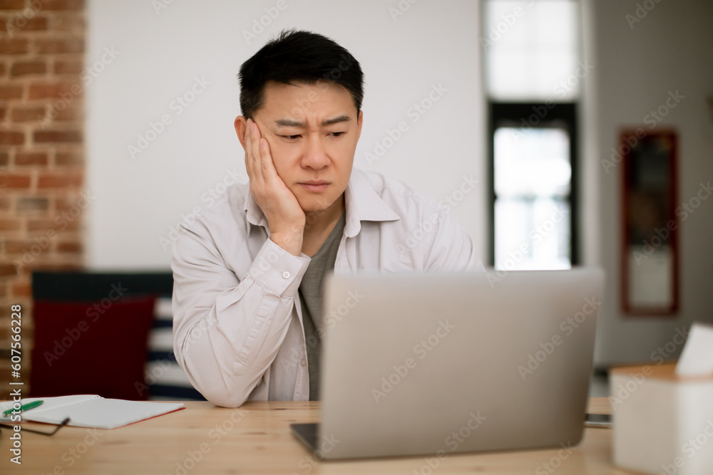 Sad tired bored middle aged man in casual looking at laptop screen, sitting at table in living room interior, free space Sad tired bored middle aged man in casual looking at laptop screen, sitting at table in living room interior, free space